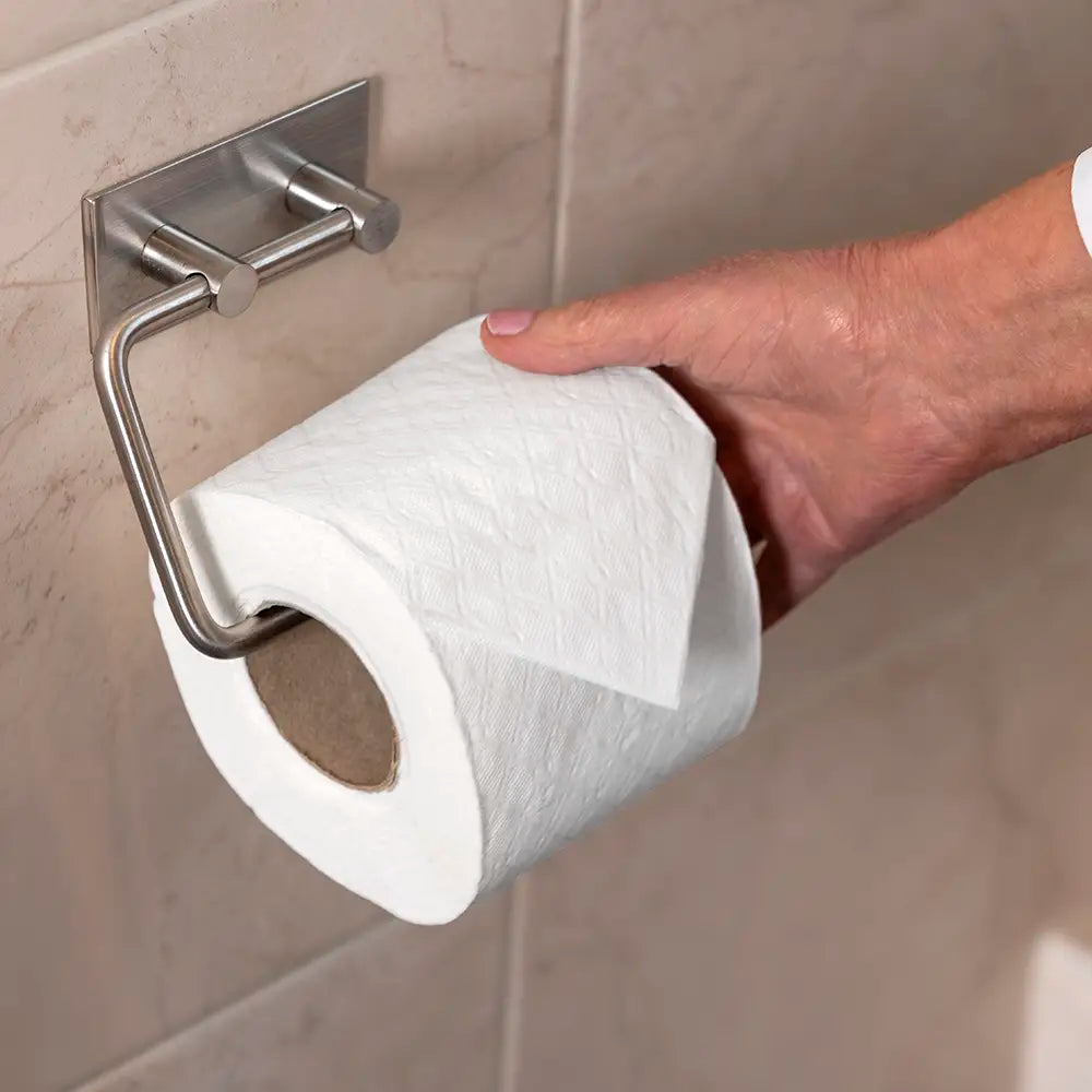 A hand holding a roll of white toilet paper against a beige tiled wall, with a brushed metal toilet paper holder mounted on the wall.