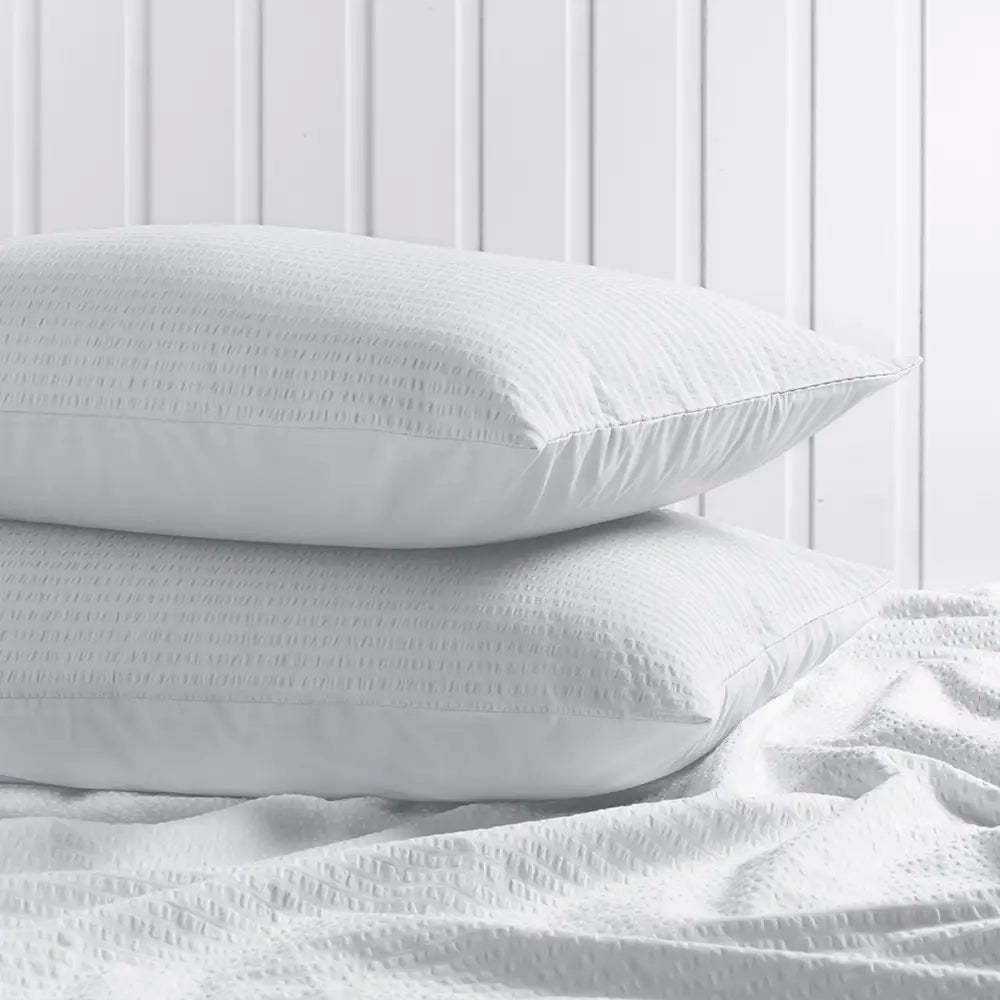 Two white textured pillows stacked on a white bed with matching white bedding in front, set against a white paneled wall background.