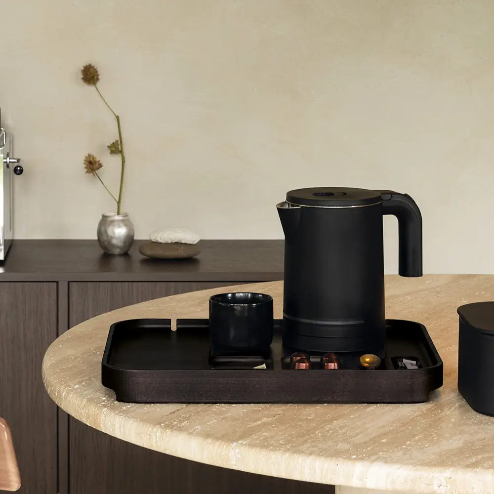 Black electric kettle on a tray with a cup and coffee pods on a kitchen counter.