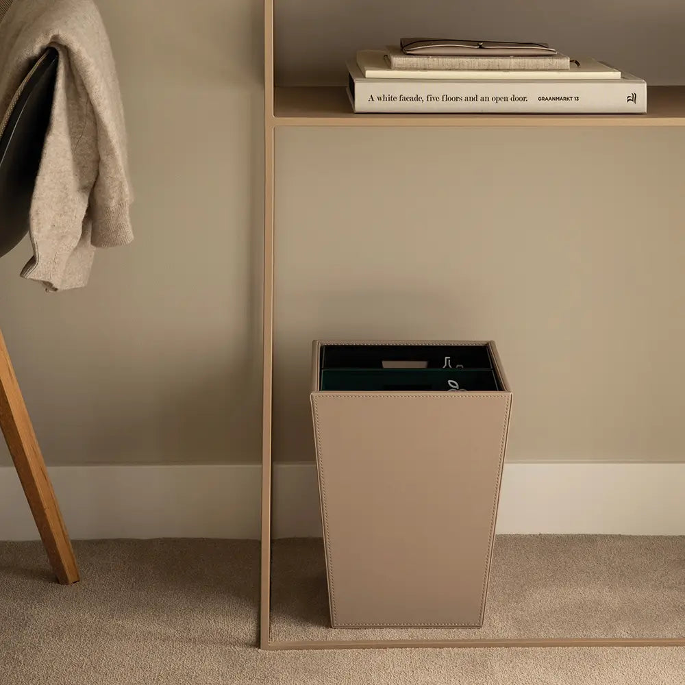 Beige trash can next to a wooden shelf with books on a beige wall.