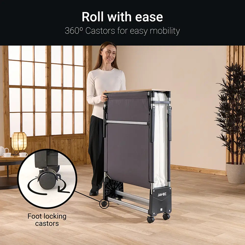 Woman pushing a folding bed with 360° castors in a room with wooden flooring and large windows.
