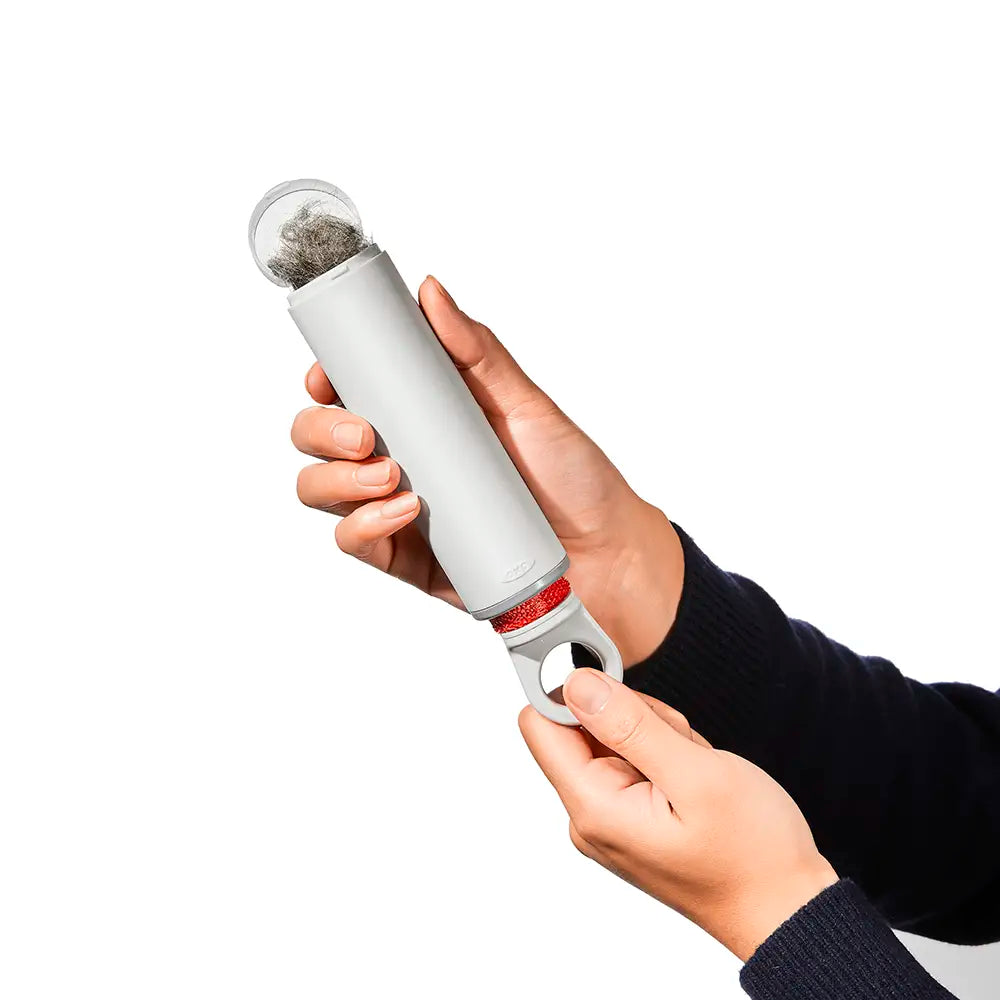 A person's hands holding a white cylindrical pet hair remover tool with open top showing trapped dark hair, against a solid white background.