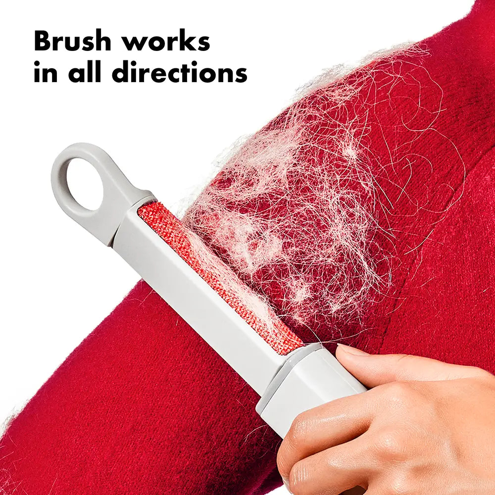 Person using a pet hair removal brush on a red fabric with text 'Brush works in all directions'.