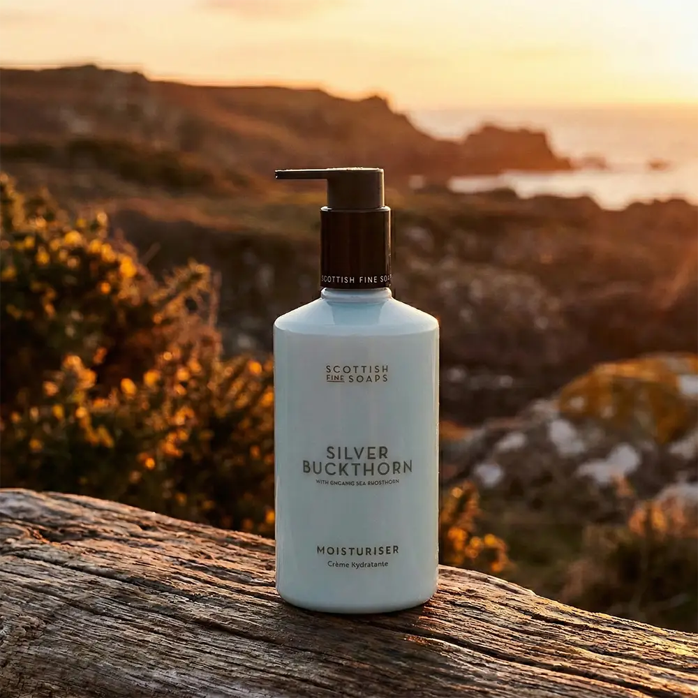 A bottle of Scottish Fine Soaps Silver Buckthorn Moisturiser stands on a weathered wooden surface with a rocky coastal landscape and ocean in the background at sunset.