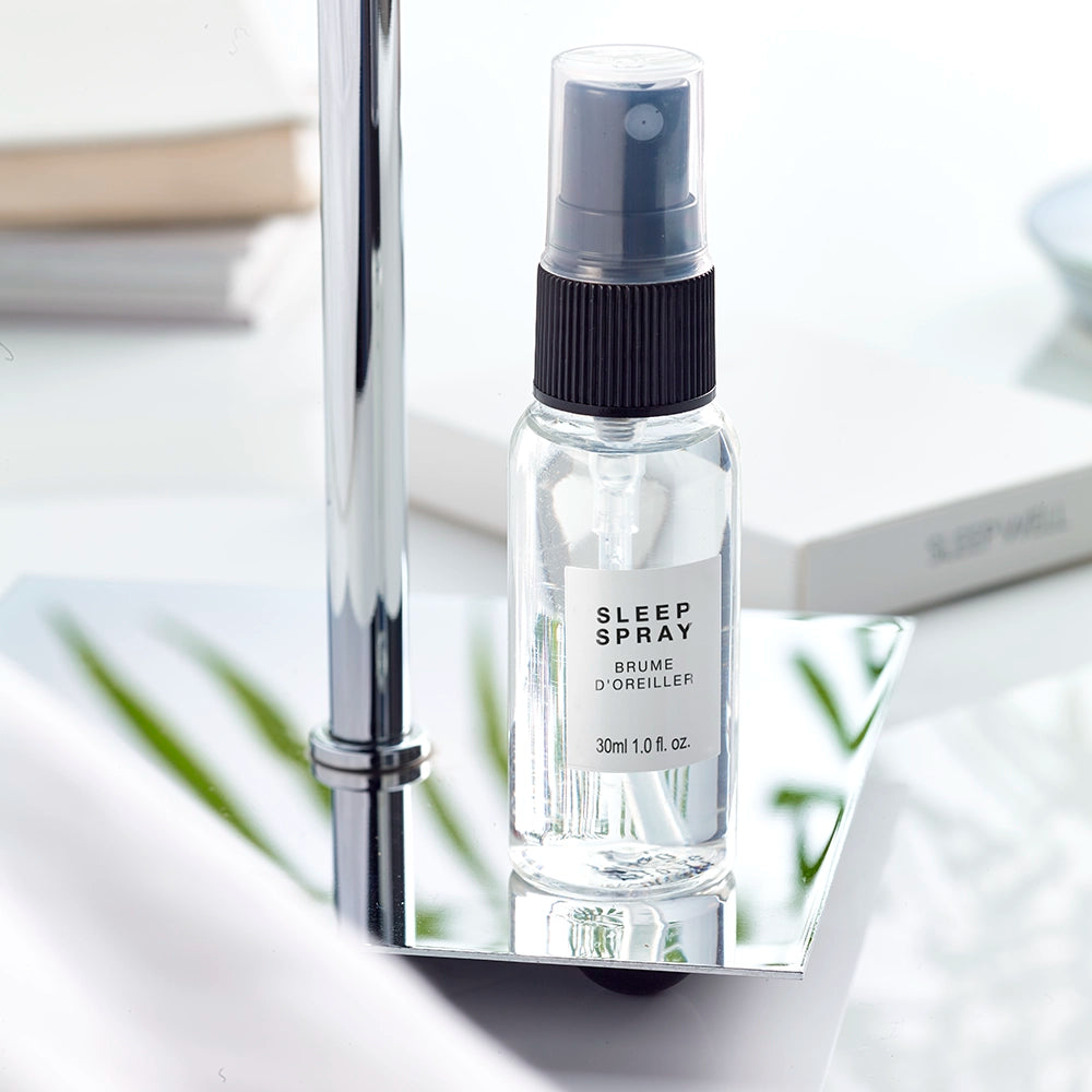 A clear spray bottle labeled 'Sleep Spray' with a black pump dispenser, placed on a silver surface, with a cap attached. The background includes blurred elements suggesting a bathroom or bedroom setting.