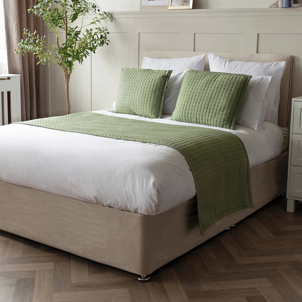 A green Crompton runner on a bed with matching pillows and a vase of flowers on the side table.