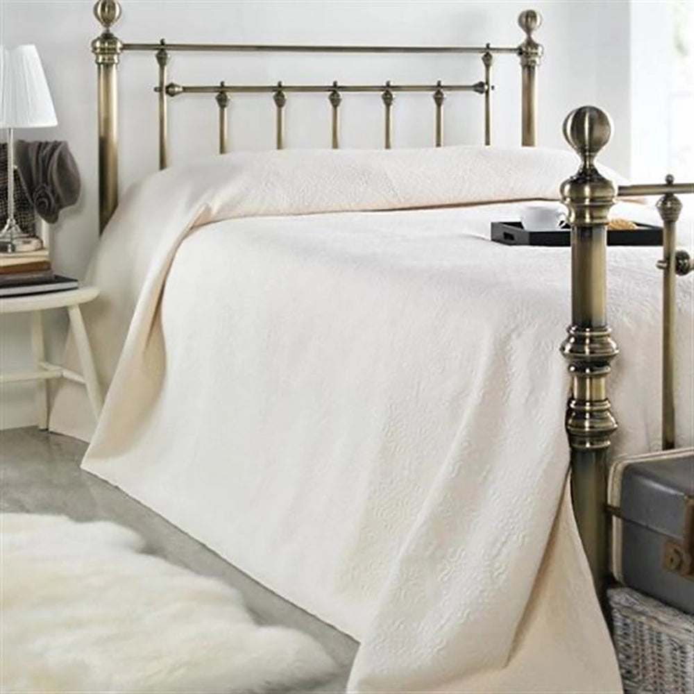 An ecru-colored jacquard patterned Brasilia bedspread displayed on a bed with ornate bedposts, in a room with a lamp and a chest of drawers in the background.