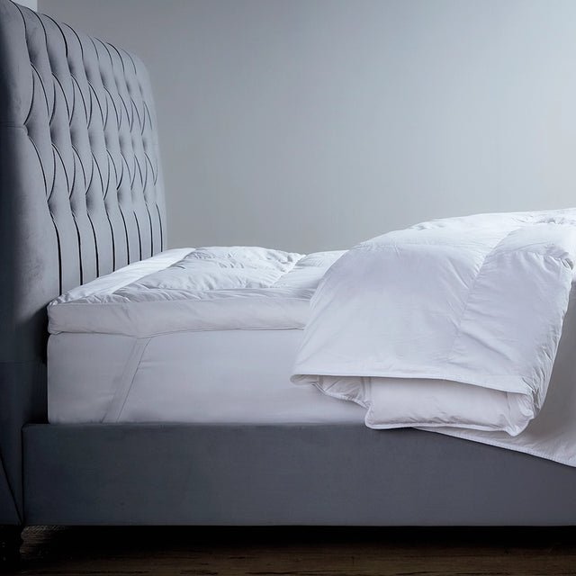 A luxury feather and down mattress topper on a bed, with a grey headboard and white bedding.