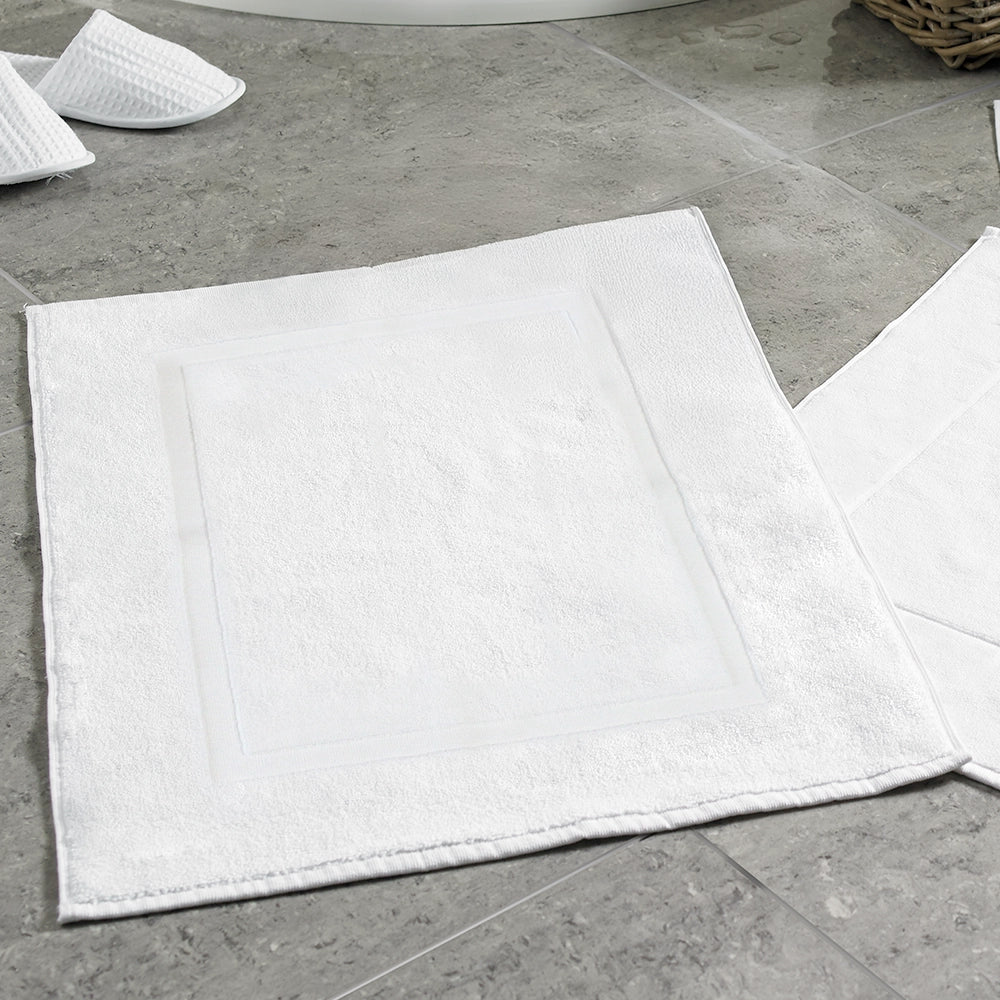 Two white 900 gsm 100% terry towling luxury cotton bath mats on a tiled floor, with one mat folded in half and the other partially visible.