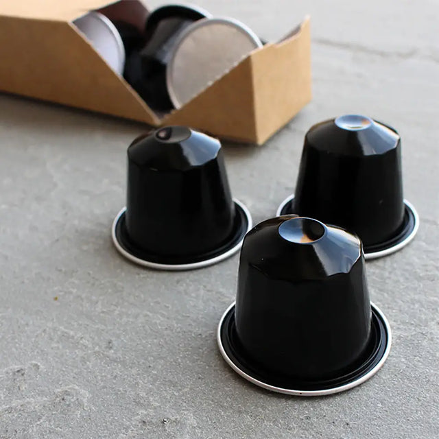 Wheatleys Fairtrade Colombian Arabica Coffee Pods - Pack of 120 -Three black coffee capsules on a concrete surface with a cardboard box in the background.