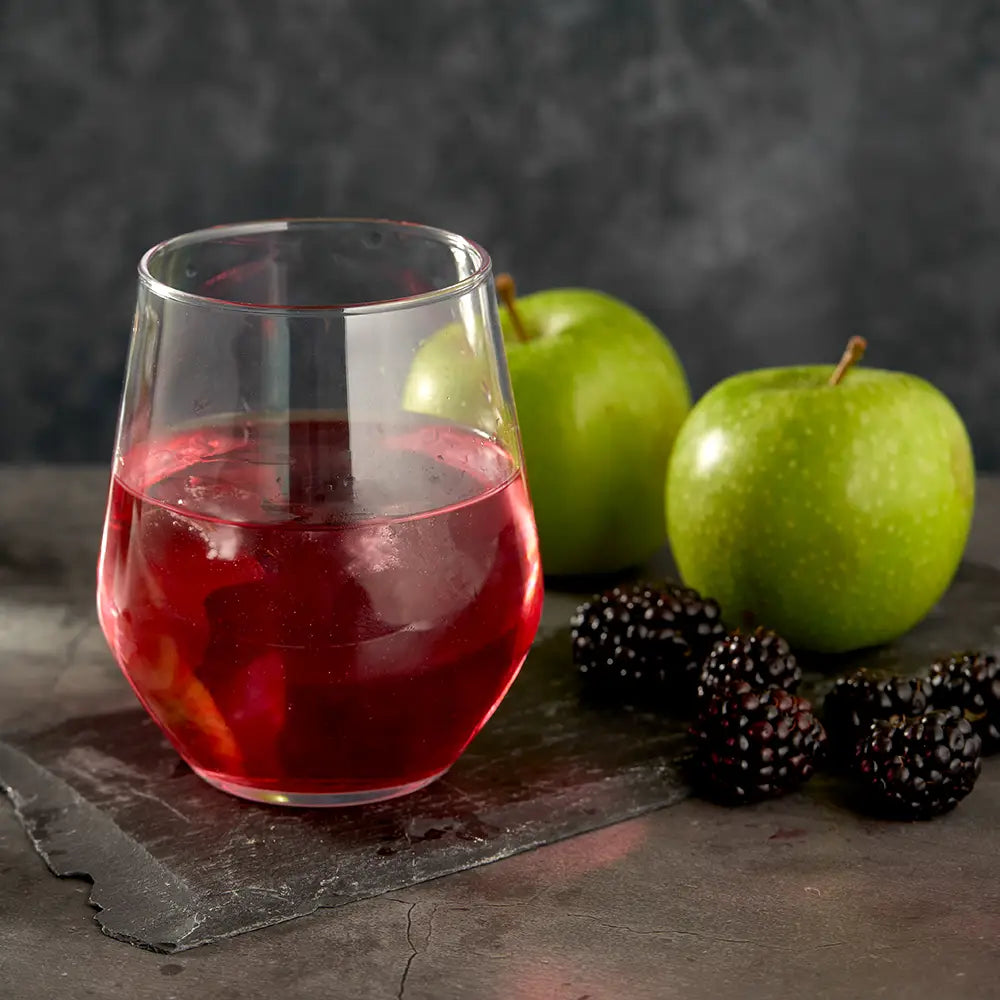 Glass of Apple and Blackcurrant Stix liquid with green apples and blackberries on a dark surface