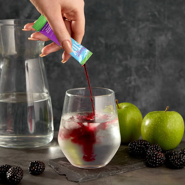 Hand pouring Apple and Blackcurrant Stix into a glass with ice, surrounded by apples and berries on a dark surface.