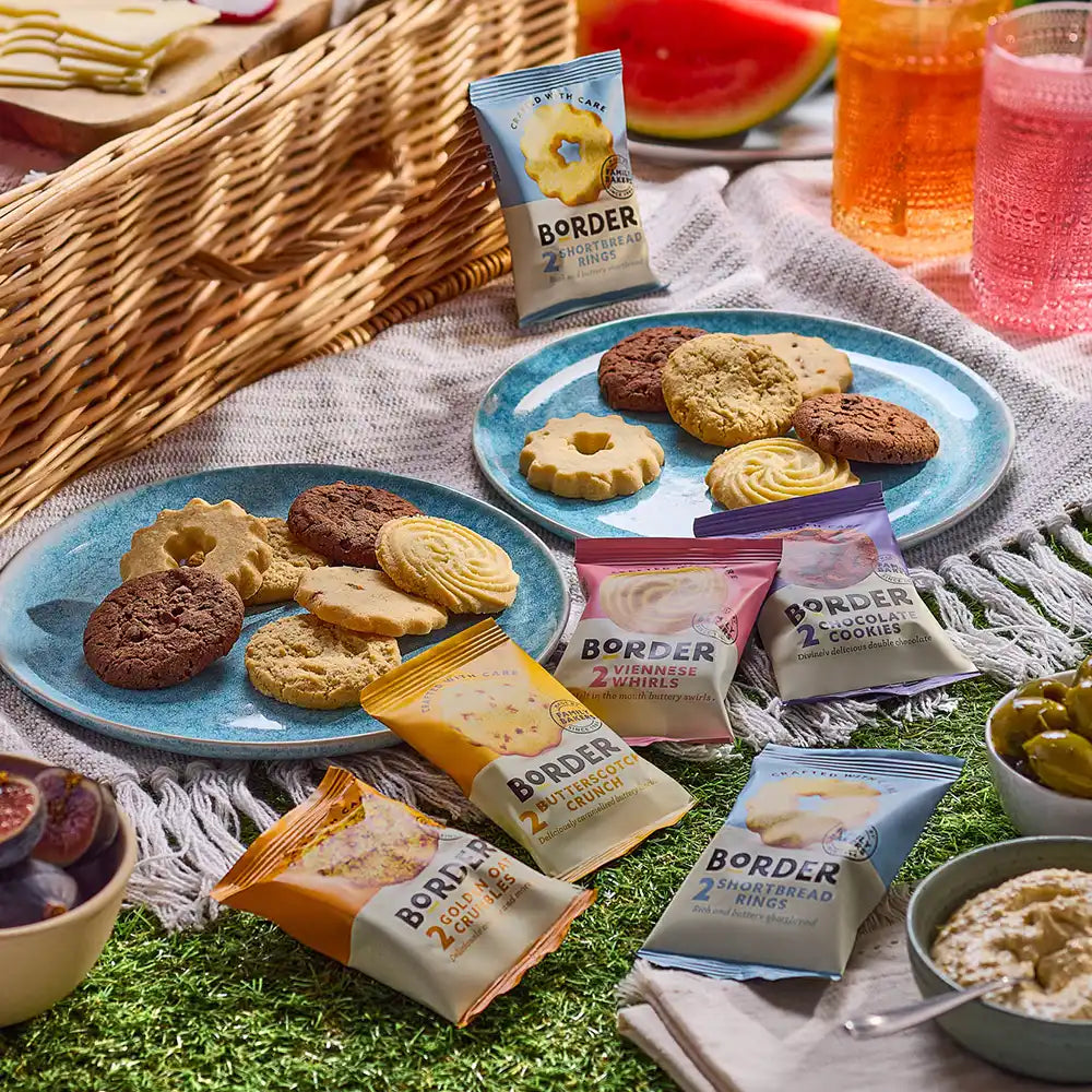 Outdoor picnic scene with plates of cookies, Border Biscuits packages, and drinks on a blanket.