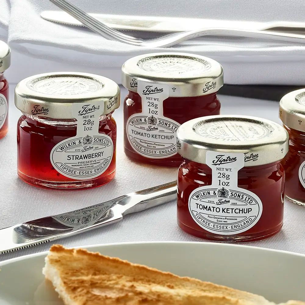 Jars of Wilkin & Sons jam and ketchup on a table with bread and a knife.