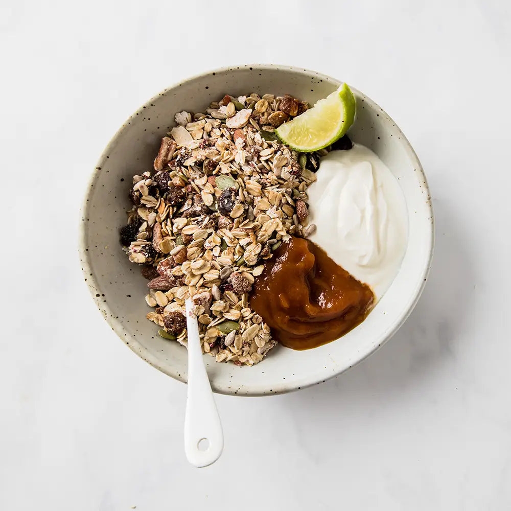 Bowl of Dorset Cereals Simply Fruity Muesli bowl of granola with yogurt, caramel, and a lime wedge on a white background
