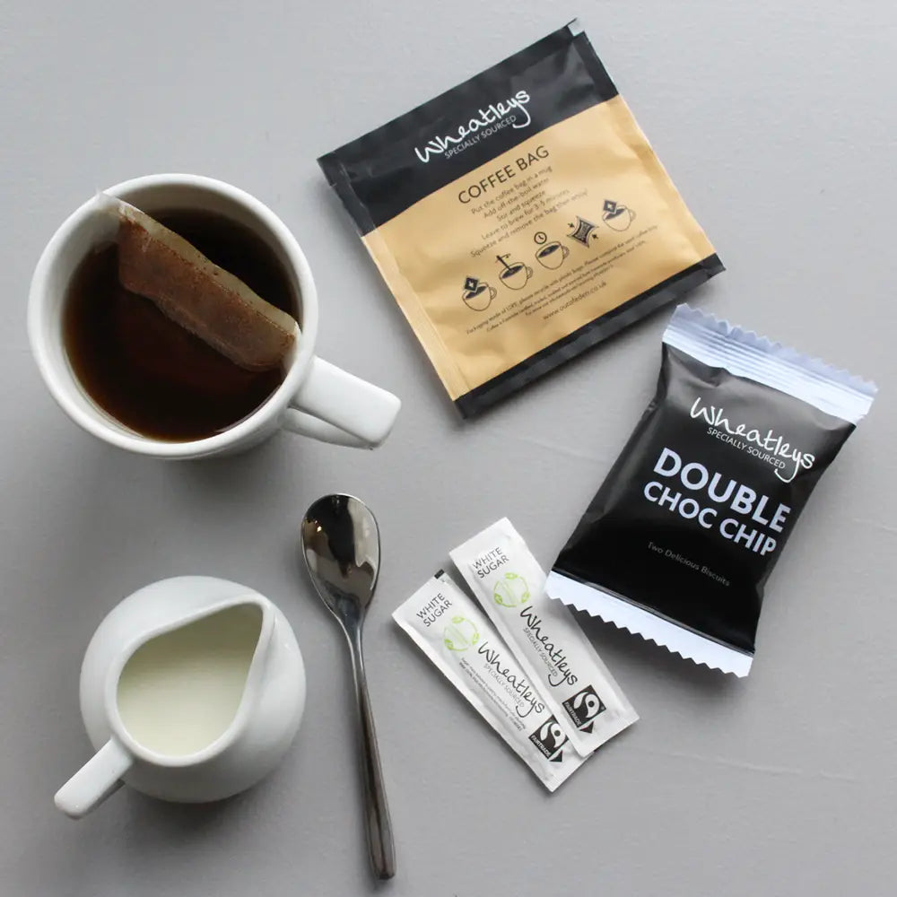 Wheatears coffee bag, double choc chip cookie mix packet, and a cup of coffee on a gray surface.