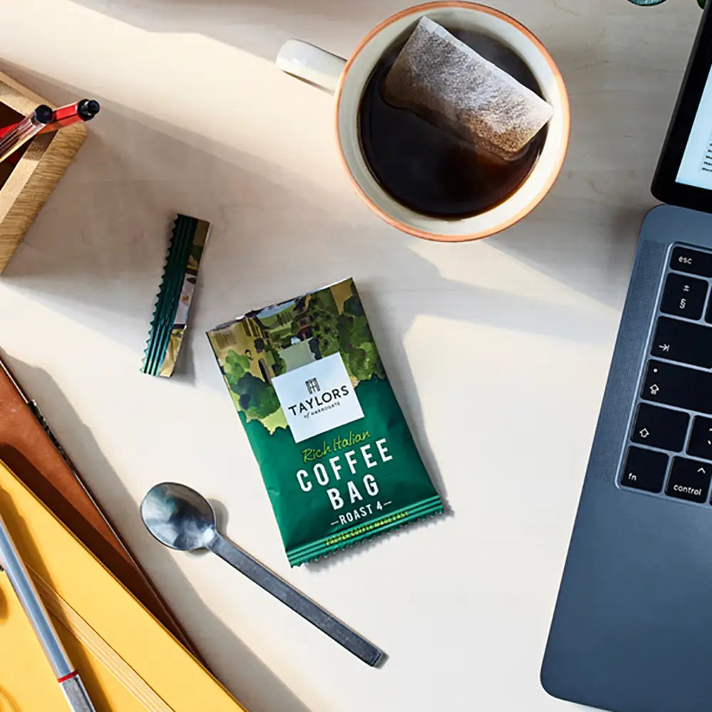 Taylor's coffee bag on a desk with a cup of coffee, laptop, and other items.