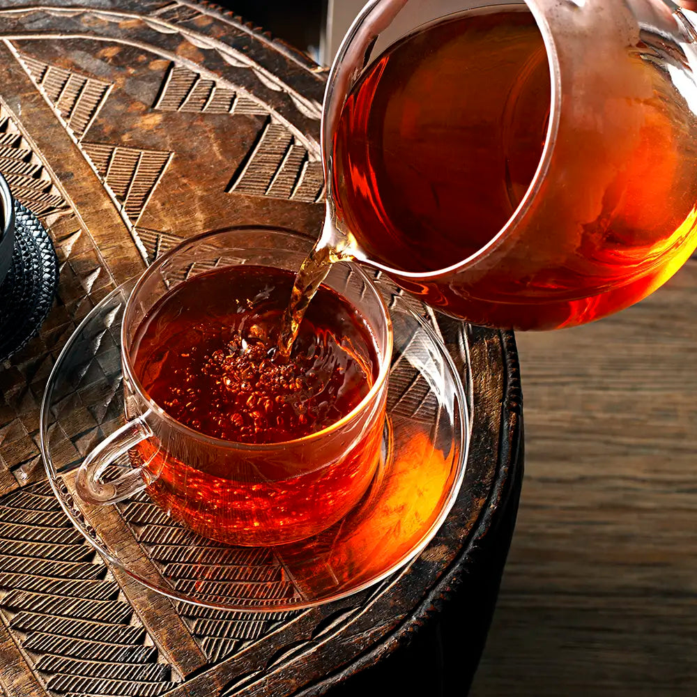 A glass teapot pours freshly brewed Birchall Cederberg Redbush tea into a glass tea cup on an ornate carved wooden table.
