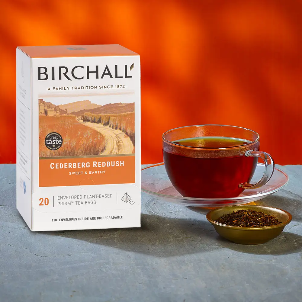 A box of 20 Birchall Redbush Tea Bags next to a cup of brewed tea and ramekin of reddish tea leaves