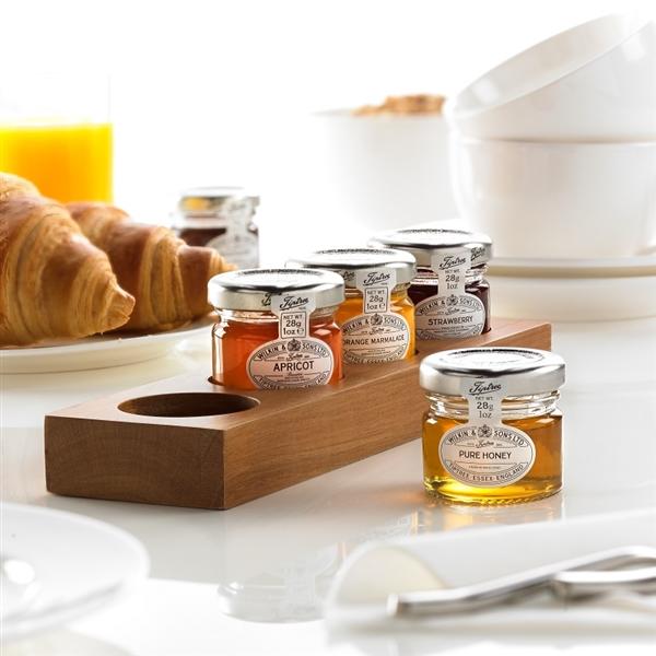 A wooden jam pot presenter with several glass jars filled with honey, placed on a white surface with a breakfast setting in the background, including a croissant and a bowl of cereal.