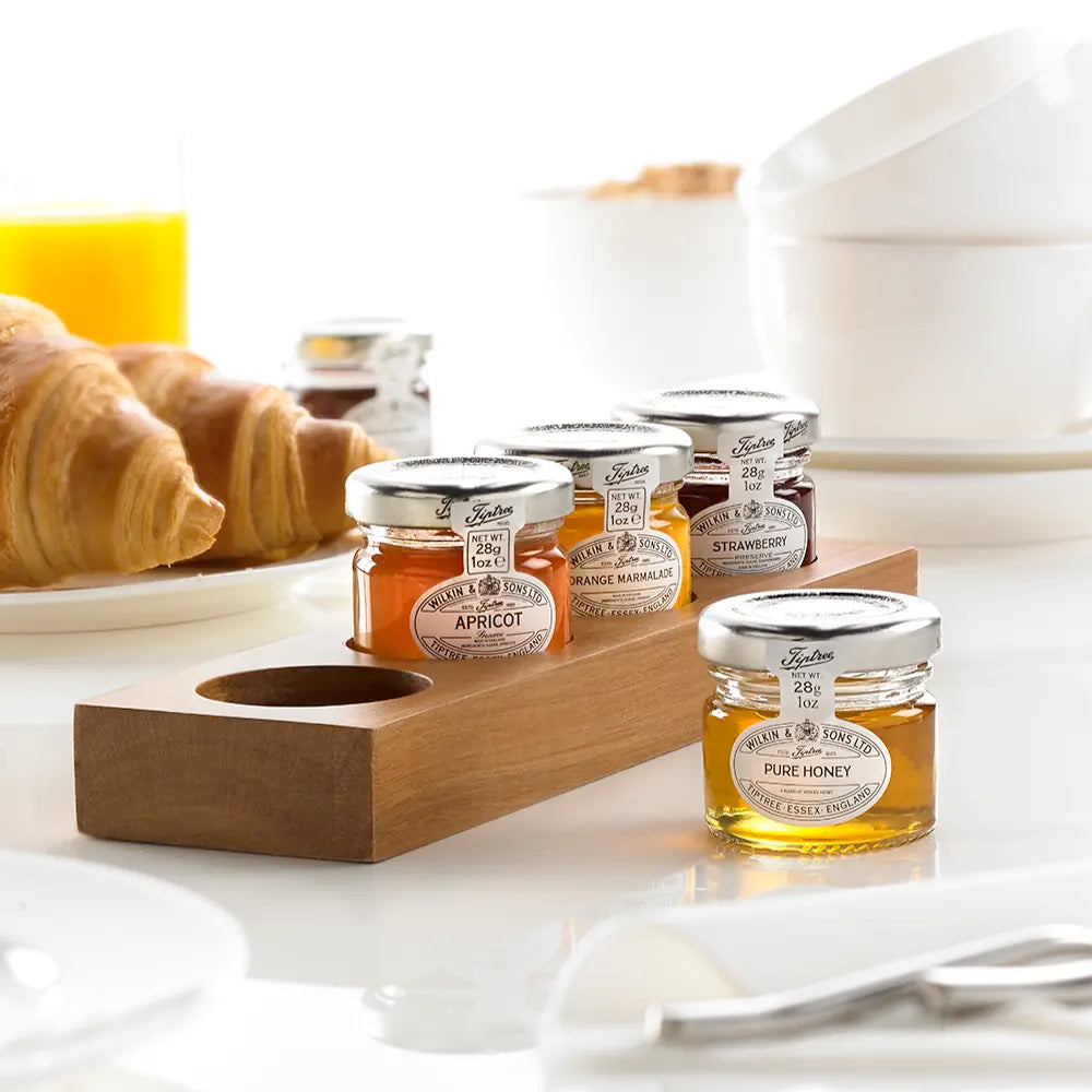 A wooden jam pot presenter with several glass jars filled with honey, placed on a white surface with a breakfast setting in the background, including a croissant and a bowl of cereal.