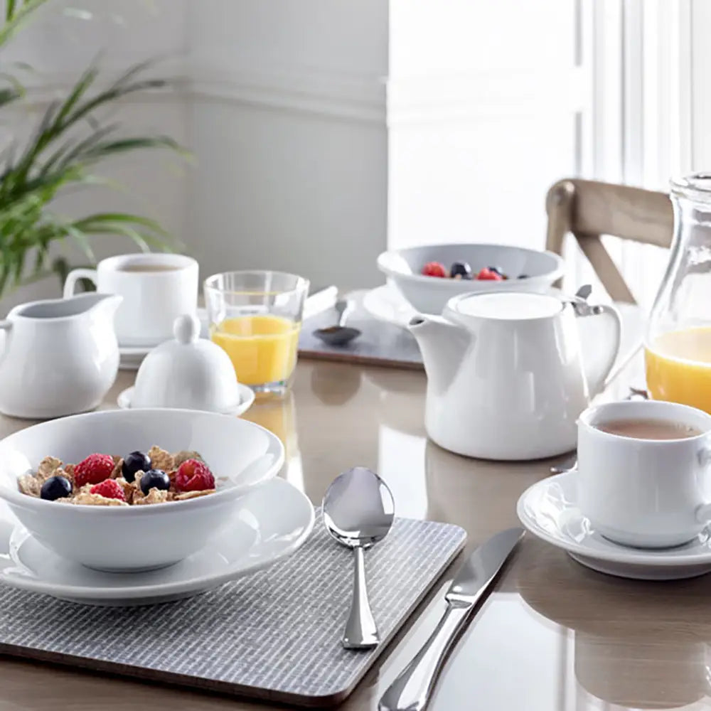 Breakfast setting with white dishes, fruit bowl, and teacup on a wooden table with Heat Resistant Melamine Placemat