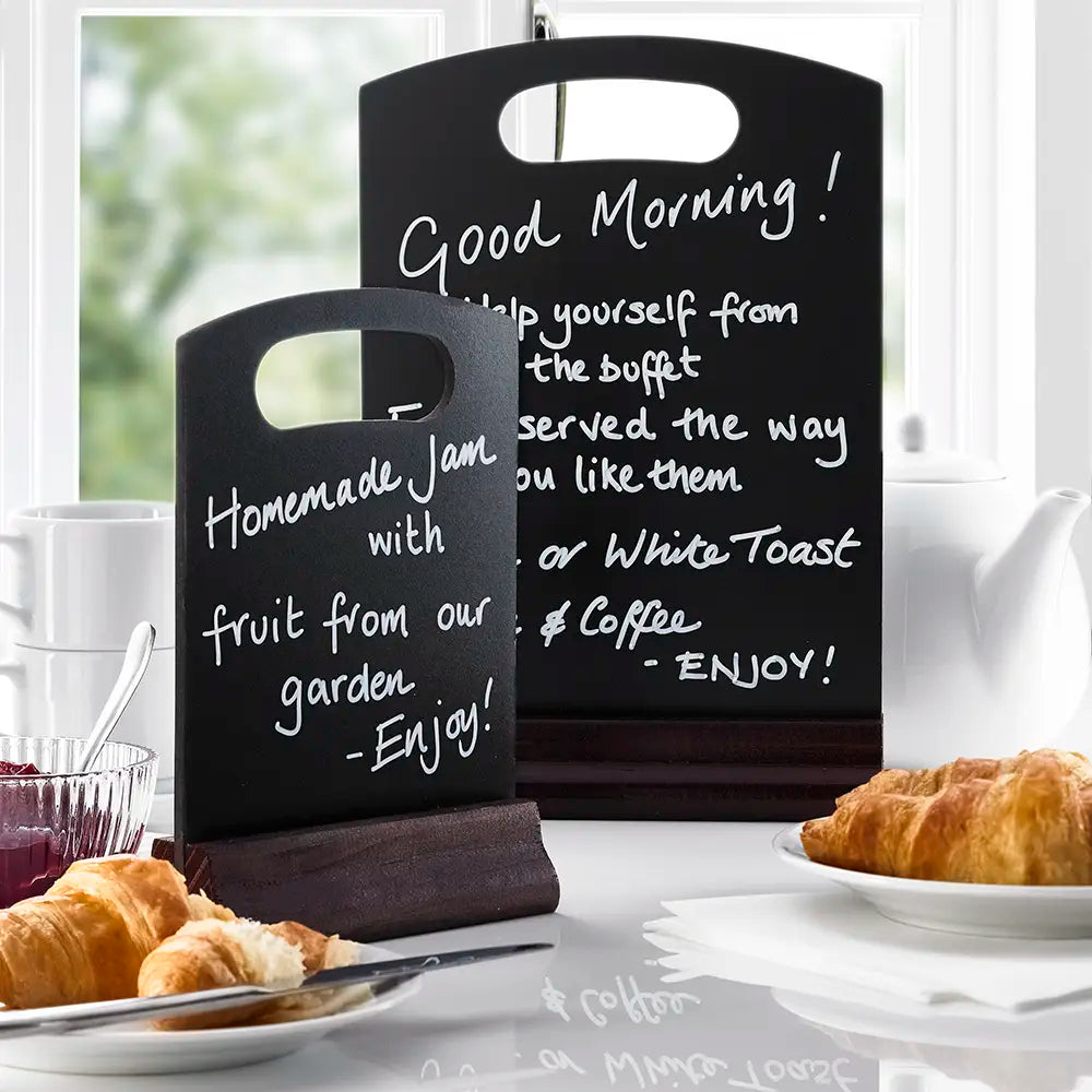 Two blackboard menus with white writing on a breakfast table with pastries and coffee.