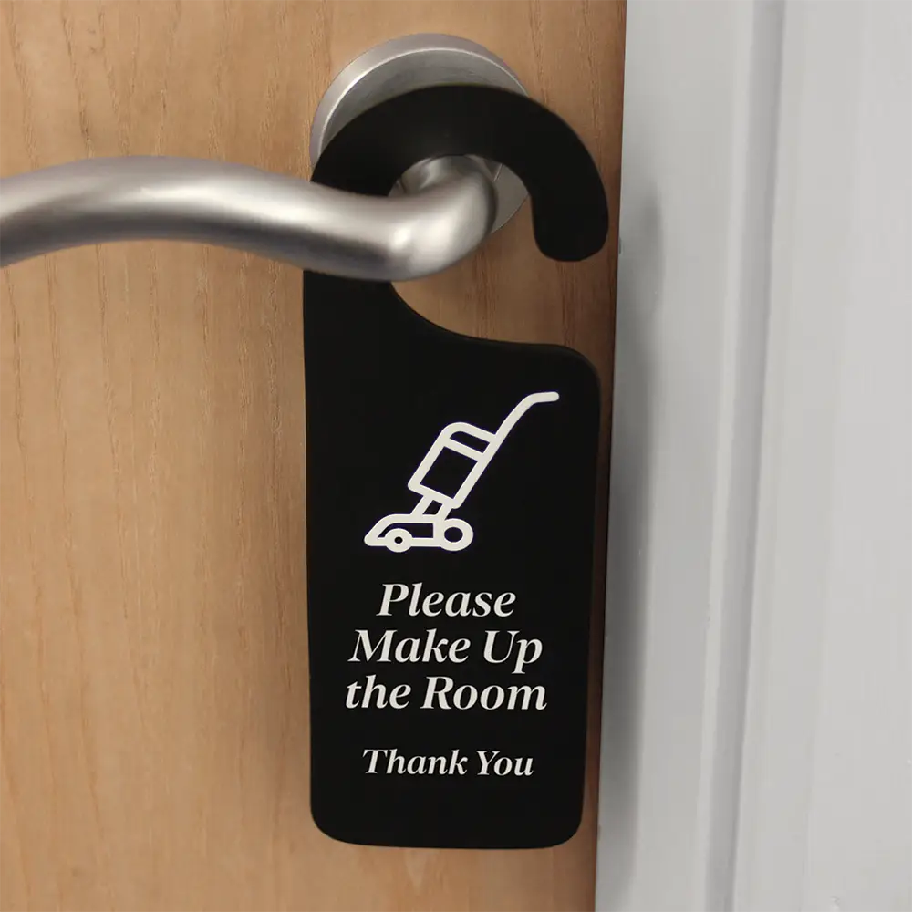 A black 'Do Not Disturb' door hanger, back of the item is displayed with text saying 'Please make up room' with white text, hanging on a door handle.