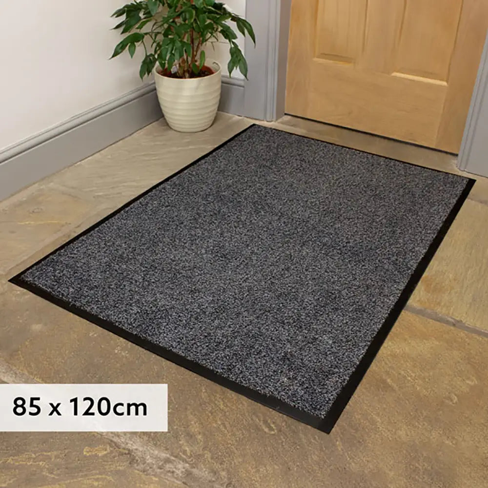Gray doormat on a stone floor with a plant and wooden door in the background