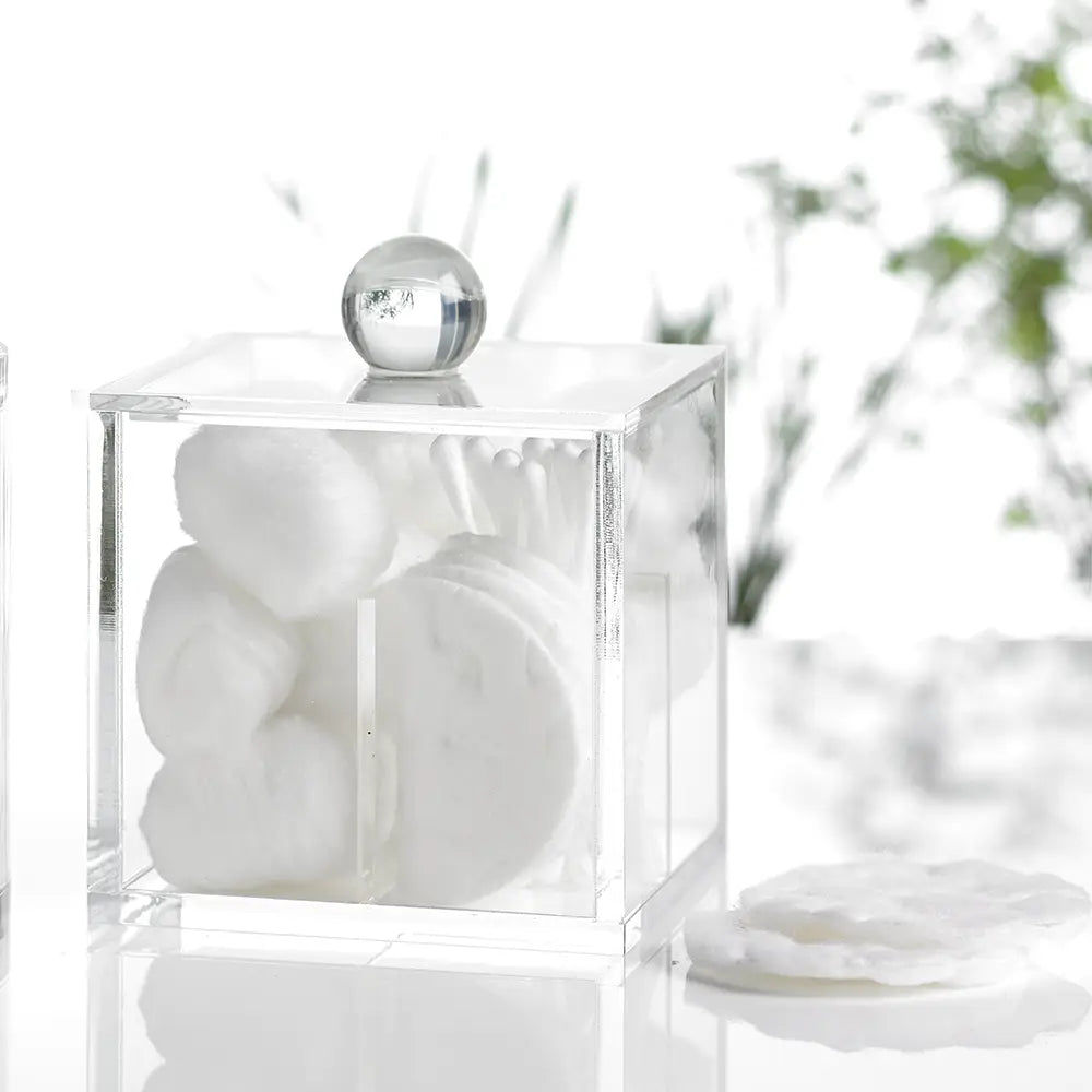 Clear acrylic cube container holding cotton wool balls, pads, cotton buds on a blurred natural background