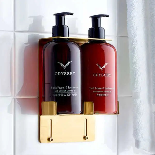 Two bottles of Odyssey shampoo and conditioner on a gold brackets against a tiled bathroom wall.