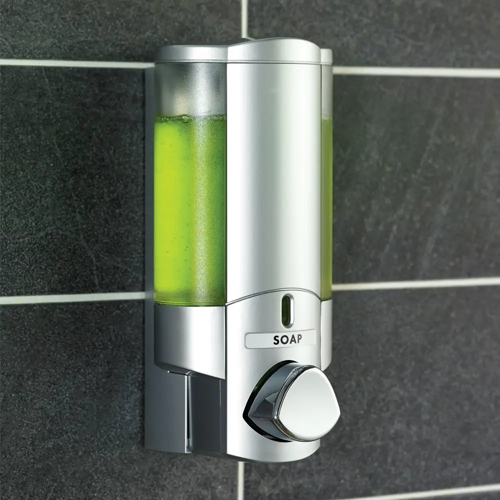 Wall-mounted soap dispenser with green soap inside, against a tiled bathroom wall.