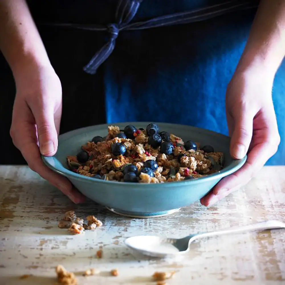 Person holding a blue bowl of Dorset Cereals Simply Delicious Muesli Portion Packs granola with blueberries on a wooden table