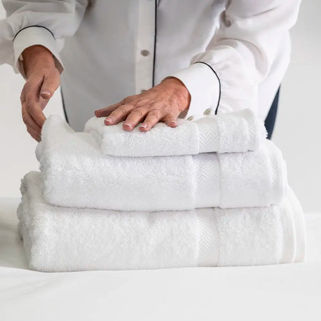 A stack of three folded white towels, including a smaller hand towel on top, with a person in a white shirt adjusting them against a clean white background.