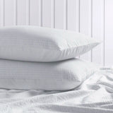 Two white textured pillows stacked on a white bed with matching white bedding in front, set against a white paneled wall background.