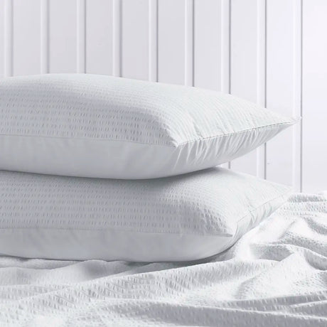 Two white textured pillows stacked on a white bed with matching white bedding in front, set against a white paneled wall background.