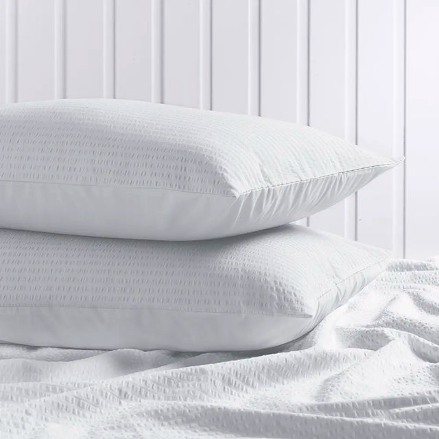 Two white textured pillows stacked on a white bed with matching white bedding in front, set against a white paneled wall background.