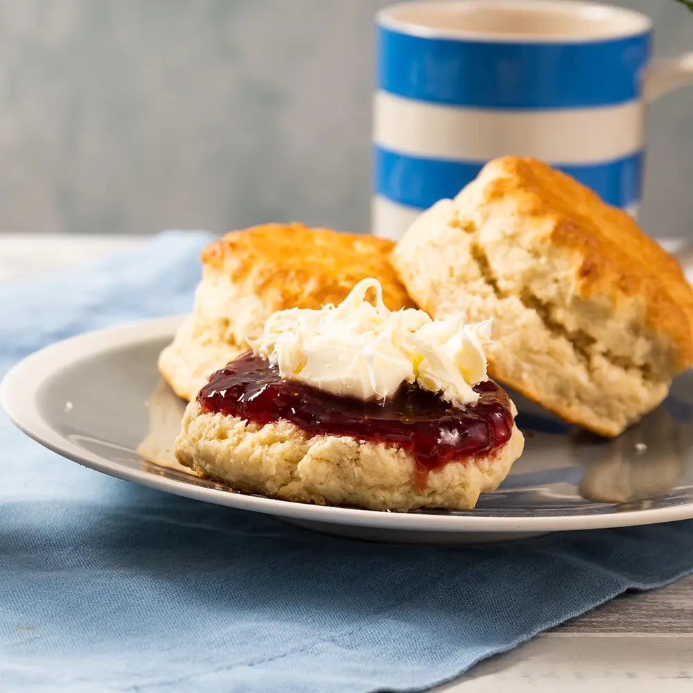 Scone with jam and cream on a plate with a blue and white striped mug in the background.