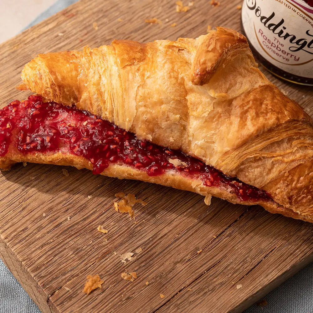 Croissant with raspberry jam on a wooden board, with a jar of raspberry jam in the background.