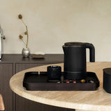 Black electric kettle on a tray with a cup and coffee pods on a kitchen counter.