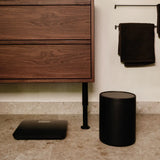 Black digital scale on a bathroom floor next to a wooden cabinet and towel rack with black towels.