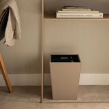 Beige trash can next to a wooden shelf with books on a beige wall.