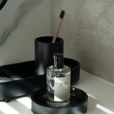 Bottle of perfume on a black tray with a toothbrush holder in the background.