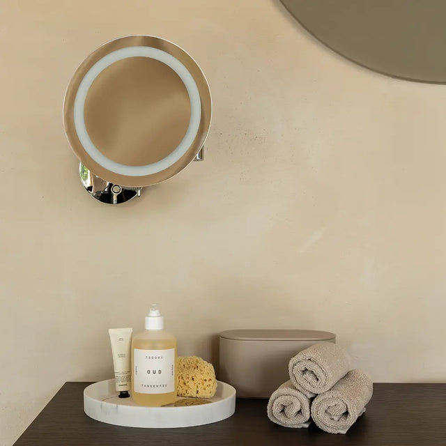 Round mirror with LED light on a bathroom counter with toiletries and towels.
