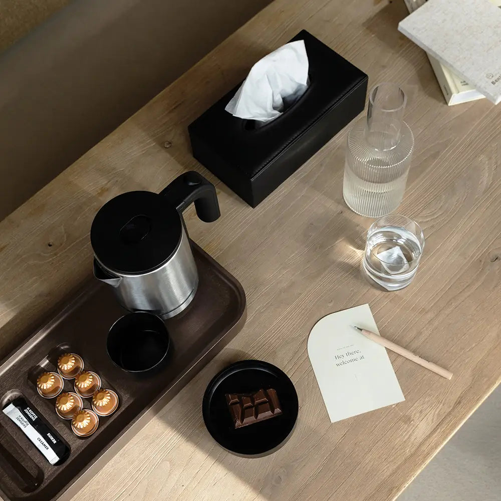 Wooden table with coffee-making items on a brown tray, a glass of water, and a tissue box