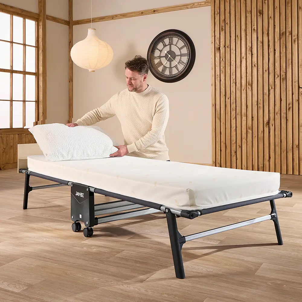 Man adjusting a mattress on a bed frame in a room with wooden walls and a clock.
