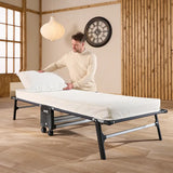 Man adjusting a mattress on a bed frame in a room with wooden walls and a clock.
