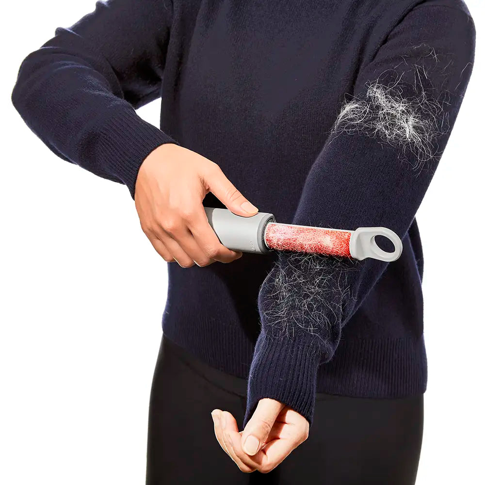 A person wearing a dark navy sweater uses a lint roller on their sleeve to remove visible white lint and hair. The person is cropped from the chest down against a plain white background.