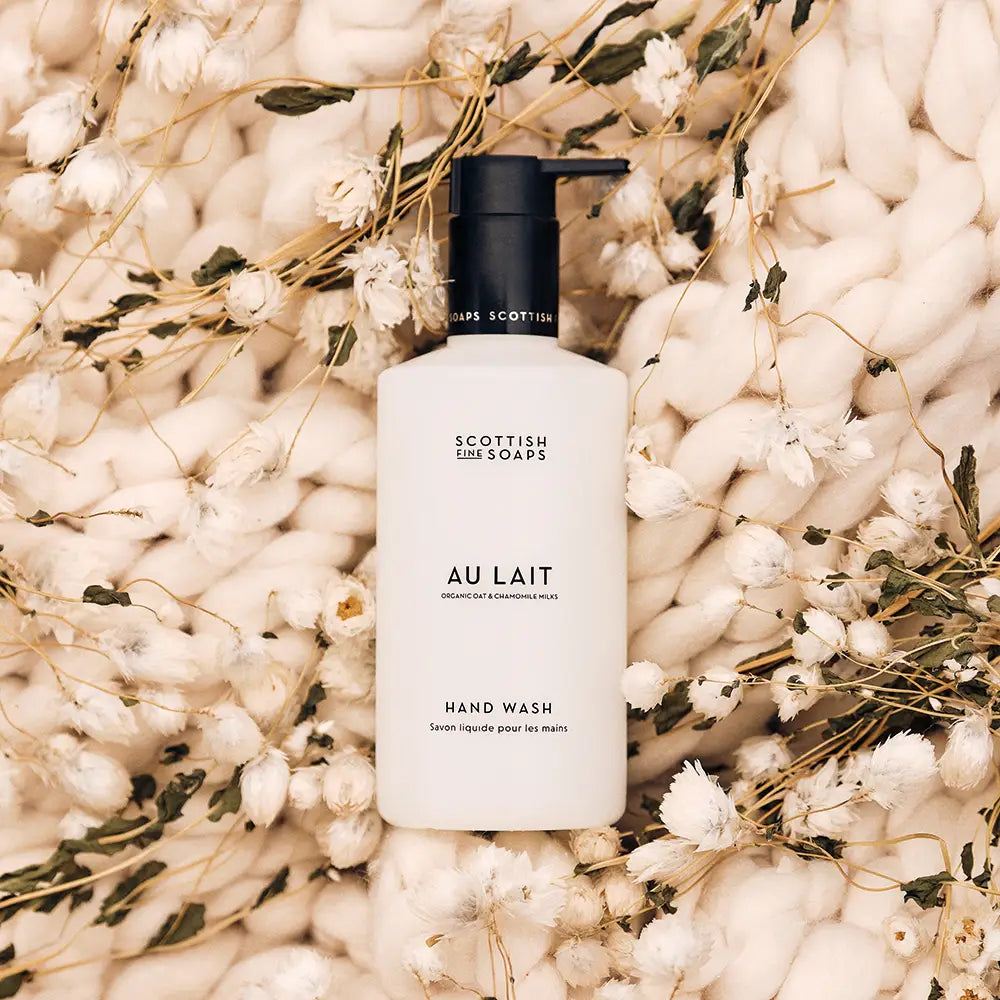 A bottle of Scottish Cosmetics Au Lait Hand Wash, featuring a matte white bottle and black pump, styled on a bed of soft cream knit fabric and dried white flowers.
