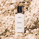 A bottle of Scottish Cosmetics Au Lait Hand Wash, featuring a matte white bottle and black pump, styled on a bed of soft cream knit fabric and dried white flowers.
